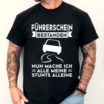 Design eines T-Shirts mit der Aufschrift "Führerschein bestanden, nun mache ich alle meine Stunts alleine" und einer Illustration eines Mannes.